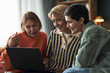 © DragonImages - Three middle aged Caucasian women sitting closely together using laptop, smiling and interacting, demonstrating friendship and connection in casual indoor setting