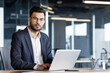 © Liubomir - A confident businessman in a navy suit and glasses, working on a laptop in a modern office setting.