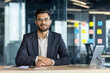 © Liubomir - A smiling businessman in a modern office setting, seated at a desk, looking directly at the camera.