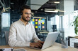 © Liubomir - A smiling man with glasses and a headset works on a laptop at an office desk. He appears to be engaged in a video call.