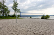 © Evgeniia - Sandy beach volleyball court set along a serene lakeshore, surrounded by lush green lawn and bushes. Inviting scene for outdoor sports, summer leisure, and an active, healthy lifestyle