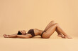 © Anastasiya - Slim Black woman in brown underwear lying on back on beige floor, arms stretched above head, legs bent, relaxed and serene in studio wellness concept.