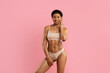© Anastasiya - Joyful young African American woman in nude underwear smiling and touching her face, posing with hand on hip against pink studio background. Confidence and body care theme.