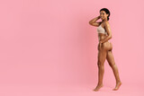 Happy African American woman in nude underwear walking confidently with hand on head, smiling against pink background. Feminine, radiant, and full of positive energy.