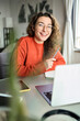 © insta_photos - Vertical portrait of happy young woman student laughing using laptop sitting at desk studying online, looking at camera. Elearning at home concept. Virtual education classes, remote work and webinars.