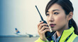 © Bakha - Female airport ground crew member in high visibility jacket communicating with walkie talkie near airplane on runway