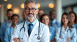 © dubogray1985 - A confident, middle-aged doctor with a friendly smile stands in the foreground, wearing a white lab coat and a stethoscope around his neck.