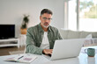 © insta_photos - Busy mature middle aged man wearing eyeglasses looking at computer technology sitting at table. Serious older man using laptop hybrid working online, elearning, browsing web, searching online at home.