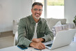© insta_photos - Portrait of happy smiling mature middle aged older professional man wearing glasses looking at camera sitting at living room table, using laptop computer working online at casual home.