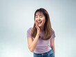 © Daniel - Asian woman in casual outfit smiling joyfully and playfully calling out with her hand near her mouth, standing against a clean light background