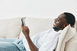 © SHOTPRIME STUDIO - Smiling African man relaxing on sofa while using smartphone, enjoying leisure time at home, casual outfit, bright and airy background for relaxation concepts