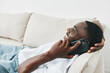 © SHOTPRIME STUDIO - Young African man happily talking on the phone while relaxing on a couch, showcasing a casual lifestyle in a light and airy living room environment