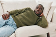 © SHOTPRIME STUDIO - A relaxed Black man in a green sweater reclining on a light colored sofa, exuding a calm and cozy vibe, perfect for lifestyle and comfort related themes