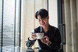 © matinee - Relaxed Asian businessman sitting by window in cafe, savoring coffee and taking a peaceful moment.