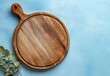 © artwiyanastudio - Round wooden cutting board on a light blue textured background, eucalyptus sprig at the left
