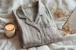 © ProStockPhoto - Folded Striped Pajama Top, Lit Candle, Jewelry on White Bedding Creates a Cozy Bedtime Aesthetic