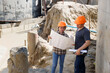 © skarie - Two construction engineers in hard hats review building plans at an active construction site. An architect and a worker discuss project details amidst a concrete construction background.