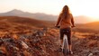 © svastix - A woman rides her bicycle on a rugged trail at sunset, surrounded by stunning mountains and warm golden hues, embodying adventure, freedom, and natural beauty in the outdoors.