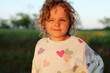 © sementsova321 - Smiling curly-haired girl in a heart-print sweatshirt stands in a sunny field, squinting in golden evening light with a playful expression.