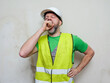 © mark_gusev - Tired workman yawning standing by a light color wall. Exhausted illegal immigrant in high visibility jacket, green shirt and white safety helmet. Working on a construction site concept.