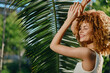 © SHOTPRIME STUDIO - smiling woman with curly hair enjoying sunlight near tropical palm leaves, radiant skin and relaxed summer atmosphere in natural outdoor setting