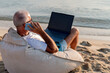 © Jelena - Mature man with laptop talking by mobile phone on beach. Business trip, watching video during preparing course work on laptop computer, sitting on the beach in sunny day. Happy work concept.