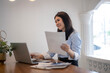 © Apichat - woman is sitting at a desk with a laptop and a stack of papers