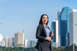 © StockPhotoRepublic - Smiling Asian businesswoman holding tablet with modern city skyline. Confident female professional outdoors in urban business district.
