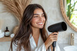 © Chatchaa - A young woman in white bathrobe blow-dries her hair at home. A radiant young woman, hair partially dried, holds a hair dryer in a bright and airy bathroom.