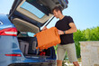 © Lazy_Bear - Man loading orange suitcase into car trunk on sunny day. Concept of preparing for summer travel, vacation or road trip with luggage