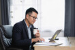 © WMSTUDIO - Professional man working on laptop with coffee cup in modern office, focusing on tasks, wearing glasses, sitting at wooden desk with papers and documents