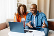 © Anastasiya - Young African American couple smiling while browsing something on laptop together, black spouses celebrating success, sitting closely on beige sofa, showing affection and focus, closeup