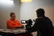 © DragonImages - Caucasian middle aged man in orange prison uniform sitting at table during interrogation, Black man in suit interviewing and recording conversation with video camera in dimly lit room