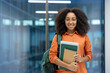 © Liubomir - A young African American woman smiles confidently, holding books and a backpack in a bright hallway.