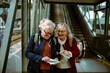 © Davor - Senior women laughing and holding travel maps at train station platform