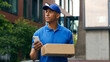 © Yuliia - Pensive African American courier man delivery guy male postman small business searching location looking at mobile phone check address smartphone deliver problem difficult in city carrying box parcel