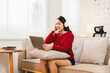 © PRIME STOCK LAB - Young woman wearing red sweater and headphones sitting on sofa with laptop, smiling and enjoying music in bright living room, relaxed and cozy atmosphere