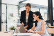 © kenchiro168 - Two Asian women working together in modern office, one standing and explaining while other sits at desk with laptop, both focused and collaborating on project