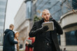 © qunica.com - Confident young man dressed in black coat, speaking on phone, writing in notebook, standing in modern urban location with blurred colleagues in background