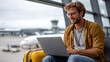 © mila103 - A man with glasses sits on a bench at an airport terminal, working on his laptop. His yellow suitcase rests beside him as he looks focused and productive in a busy travel environment.