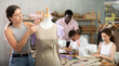 © JackF - Interested young woman adjusting and pinning fabric on mannequin during hands-on dressmaking class, while classmates sewing and preparing materials at table in background..