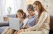 © Studio Romantic - Grandmother, mother and child girl sitting on sofa using laptop together having online video call or watching movie. Smiling happy family resting on couch at home. People and technology concept.