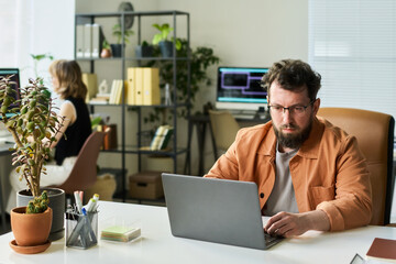 Caucasian middle aged man working on laptop developing neural network in modern office, sitting at desk with potted plant, female colleague using computer in background