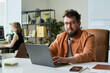 © Mediaphotos - Portrait of Caucasian young adult man sitting at desk working on laptop developing neural network, looking into camera with slight smile, office environment with blurred colleague in background