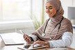 © Prostock-studio - Distance healthcare, telehealth technologies. Cheerful muslim woman doctor sitting in her cabinet, using digital tablet and mobile phone, copy space. Lady in hijab physician working online from clinic