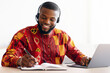 © Prostock-studio - Online Learning. Cheerful Black Man In Traditional African Clothes Study With Laptop And Headset At Home, Positive Millennial Guy In Ethnic Shirt Noting Down Information To Notepad, Free Space