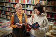 © Seventyfour - Caucasian middle aged woman and Asian young adult woman standing in bookstore reading books together, surrounded by shelves filled with books, engaging in discussion