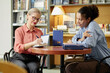 © Seventyfour - Caucasian senior woman autographing book while sitting at table with Black young adult woman, both focused on discussion in library or bookstore setting during session
