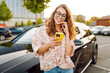 © maxbelchenko - Beautiful woman with curly hair with a phone in her hands stands near a car in a parking lot. Young female driver enjoys the weather and chats at sunset. Transport and technology concept.