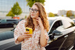 © maxbelchenko - Beautiful woman with curly hair with a phone in her hands stands near a car in a parking lot. Young female driver enjoys the weather and chats at sunset. Transport and technology concept.
